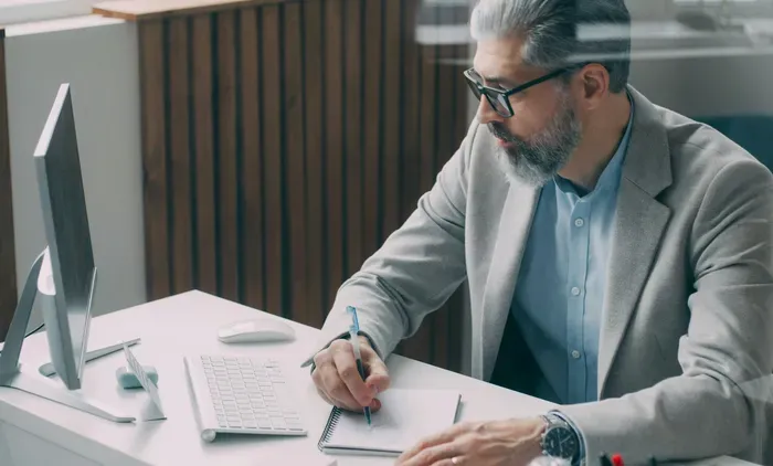 Grey-bearded professional takes notes at a modern desk with computer and keyboard, planning tasks for a company